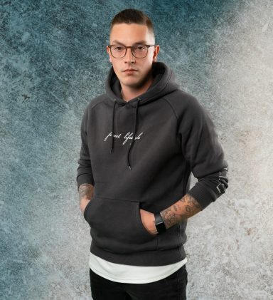 Junger Mann in einem grauen Hoodie mit Tattoo, vor einem strukturierten Hintergrund.
